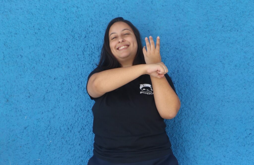 Uma jovem mulher de olhos e cabelos compridos castanhos sorri. Está com o braço esquerdo levantado e com a mão aberta; o braço direito, com a mão fechada, encosta na direito, indicando o nome baleia em Libras